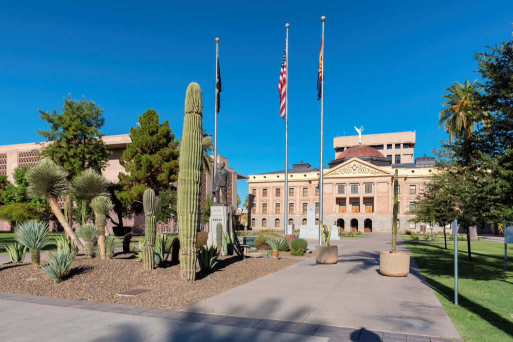 Arizona State Capitol building with saguaro cacti and desert landscaping, US and Arizona flags against blue sky
