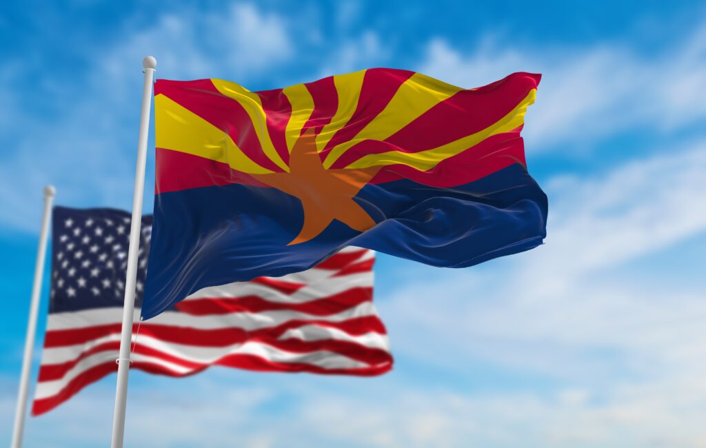 Arizona state flag waving on a pole with an American flag blurred behind against a blue sky