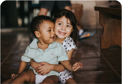 Two young children on a tiled indoor floor; older girl hugs and smiles at a toddler boy.