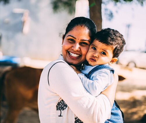 Smiling parent in white top embraces toddler outdoors under a tree