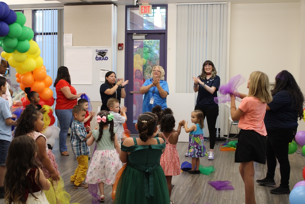 Healthy Families graduation with children and staff clapping and dancing in a community room with rainbow balloon arch
