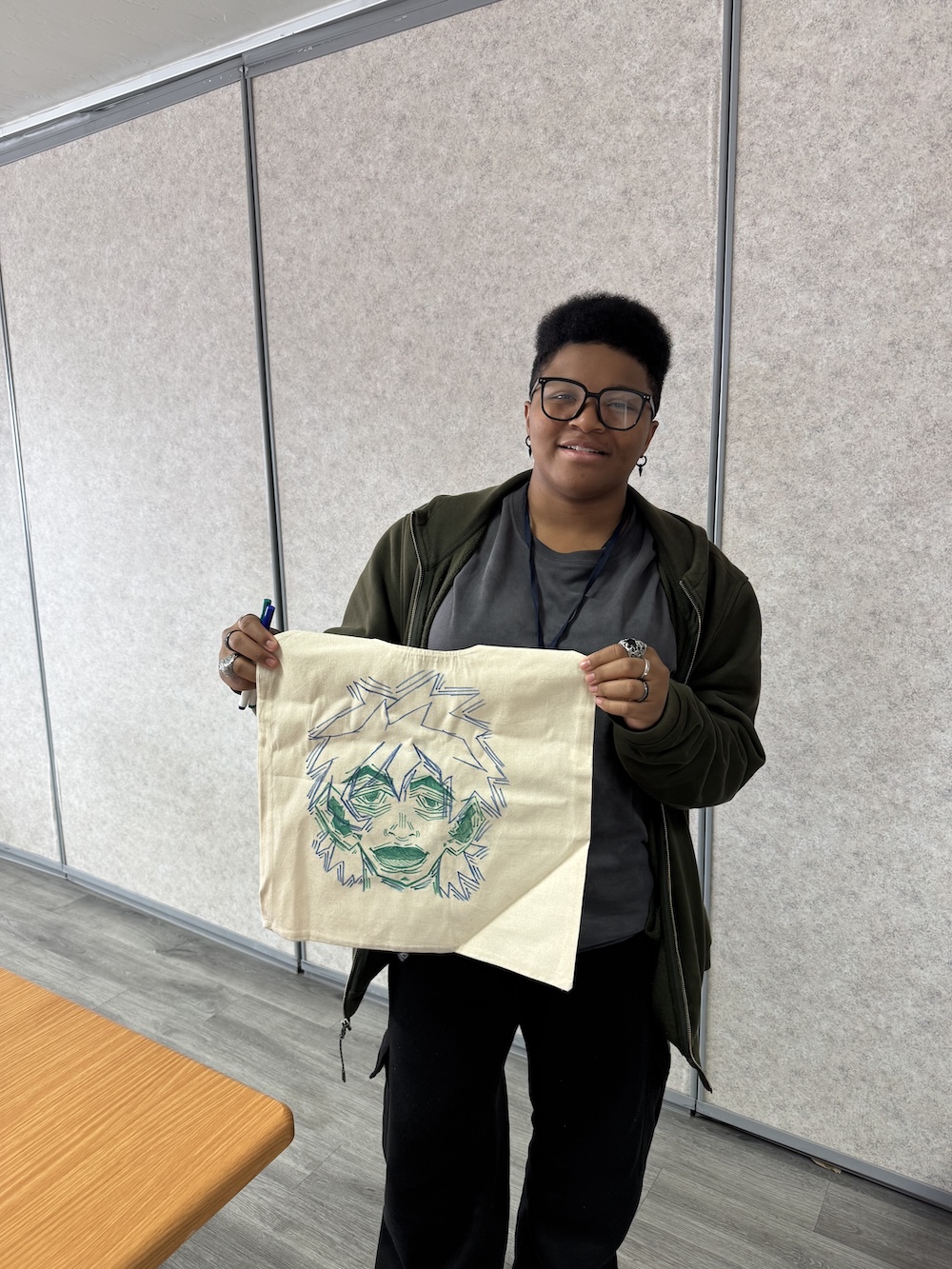 Smiling teen showing a canvas bag with a green anime-style face drawing at arts & craft day